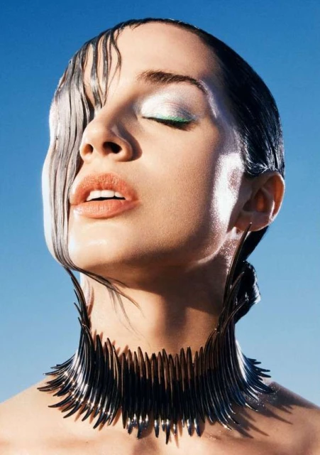 Close-up beauty shot of a woman with wet hair, closed eyes, and decorative neckpiece against a clear blue sky background.