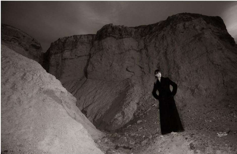 Model wearing a long black coat standing in a rocky desert landscape under an overcast sky.
