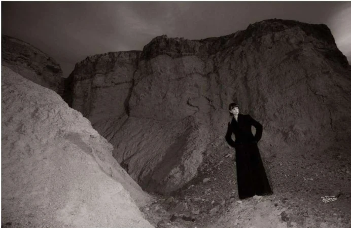 Model wearing a long black coat standing in a rocky desert landscape under an overcast sky.