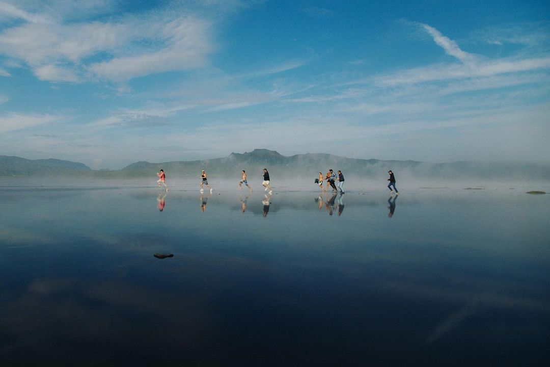 Group of people running on wet sand near the ocean with blue sky and mountainous background reflecting in water, creating a mirror effect.