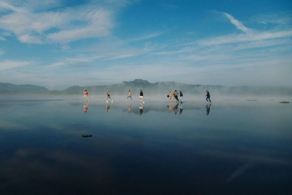 Group of people running on wet sand near the ocean with blue sky and mountainous background reflecting in water, creating a mirror effect.