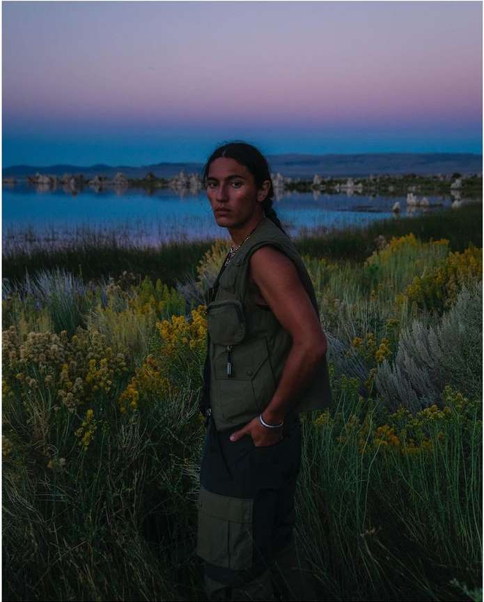 A lone man stands in an olive vest and dark pants amid a reedy field near a lake, under a dusk sky in a landscape shot.