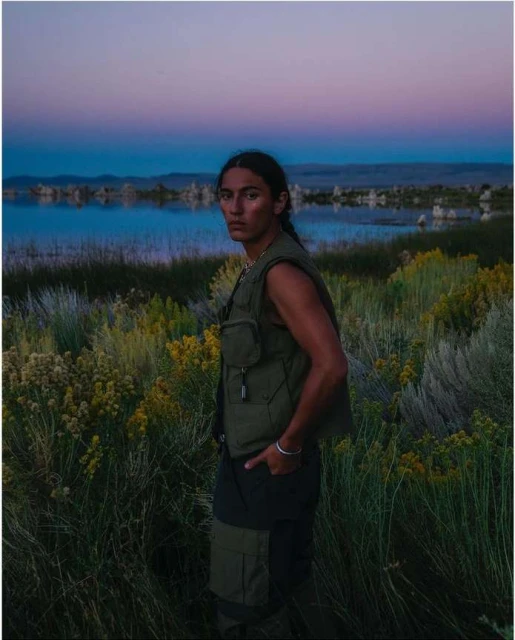 A lone man stands in an olive vest and dark pants amid a reedy field near a lake, under a dusk sky in a landscape shot.