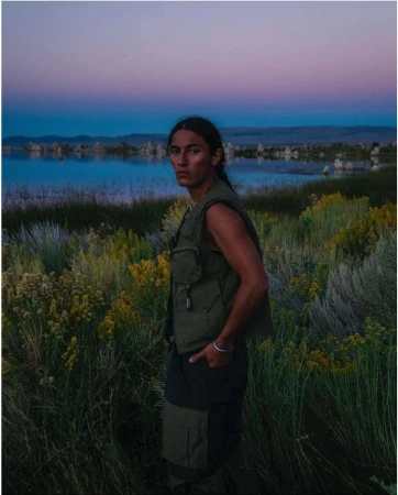 A lone man stands in an olive vest and dark pants amid a reedy field near a lake, under a dusk sky in a landscape shot.