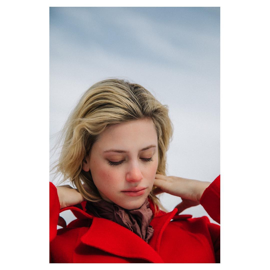 Blonde woman in red coat poses outdoors against a cloudy sky. She has her eyes closed, and her hands are behind her neck.