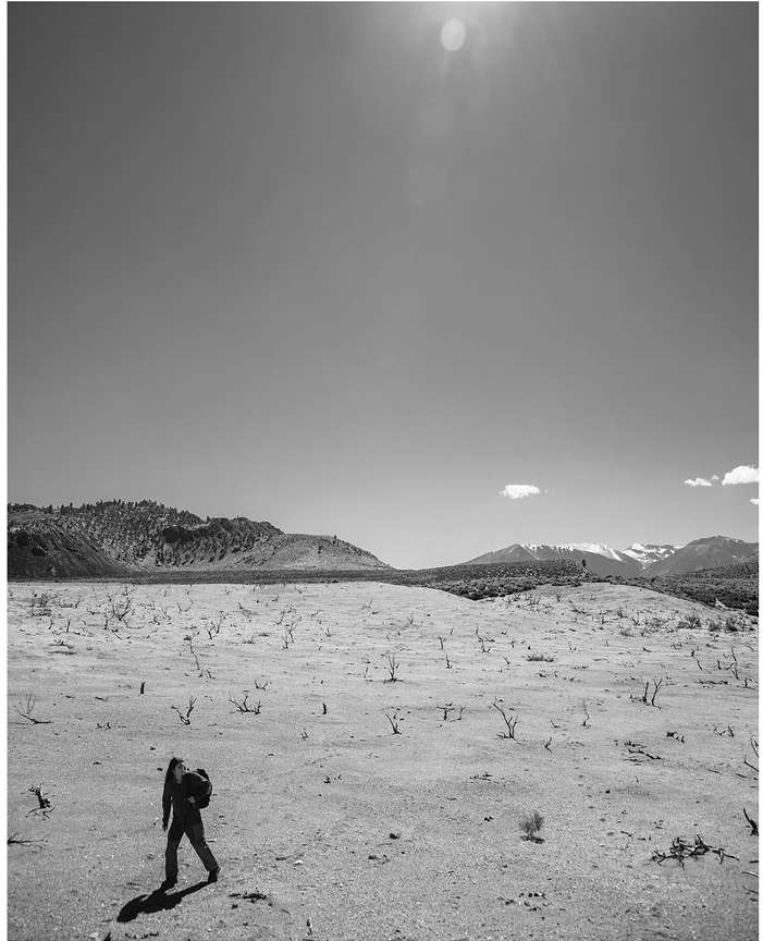 Black and white landscape shot of a person walking through a valley with hills and mountains in the background under a bright sky.