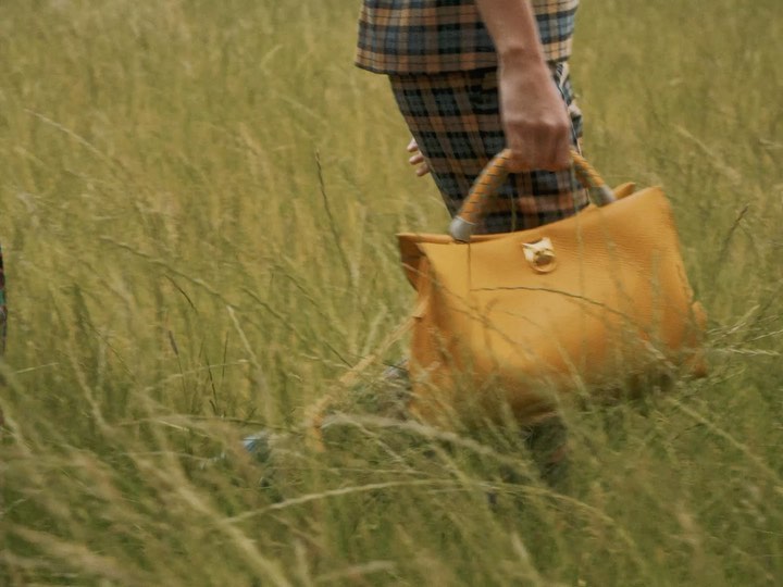 Person walking through a field carrying a large yellow handbag, wearing a plaid outfit, creating a fashion-forward visual.
