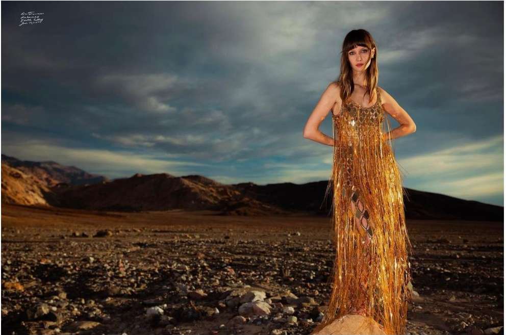 A woman in a gold dress stands in a desert landscape with mountains and a cloudy sky in this fashion editorial photo.