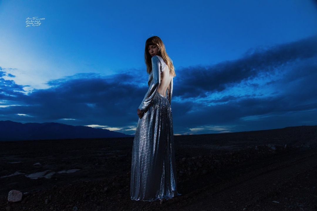 Woman in a long metallic silver gown standing outdoors under a dramatic blue twilight sky by Cole Sprouse