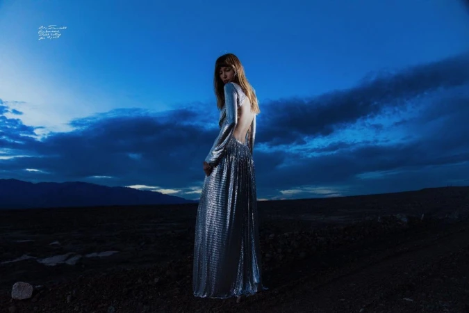 Woman in a long metallic silver gown standing outdoors under a dramatic blue twilight sky by Cole Sprouse