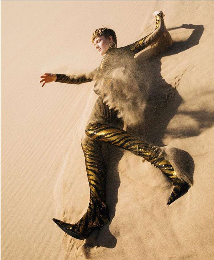 Fashion shot of a blonde woman in gold and black tiger-striped outfit posing on a sand dune with sand blowing around her.