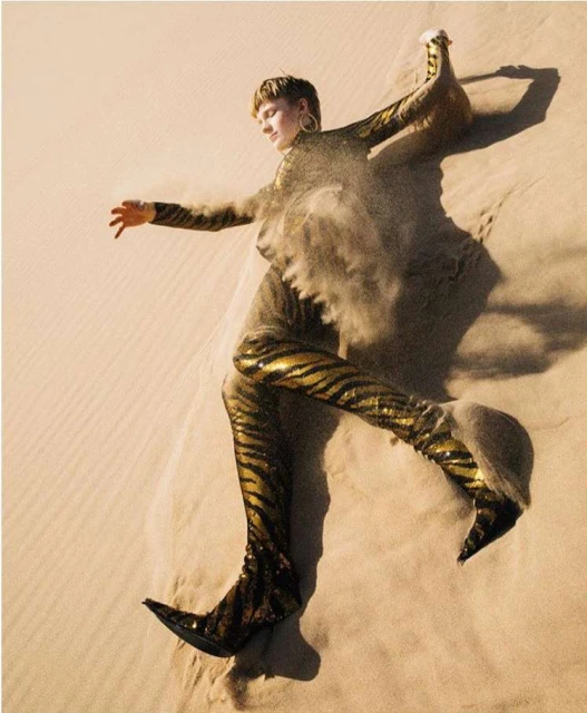 Fashion shot of a blonde woman in gold and black tiger-striped outfit posing on a sand dune with sand blowing around her.