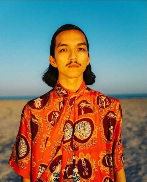 Portrait of a young adult male outside wearing an orange patterned collared shirt against a bright blue sky and tan sandy background.