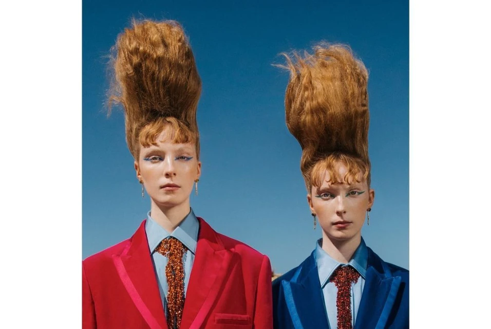 Portrait of two women with tall auburn hairstyles wearing colorful suits against a solid blue sky, striking a poised, confident pose.