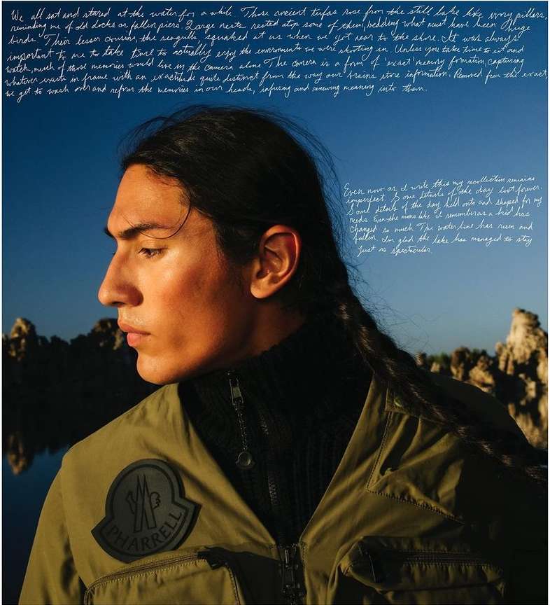 Profile portrait of an adult male with long braided hair, wearing olive green outerwear in an outdoor lakeside setting with rock formations.
