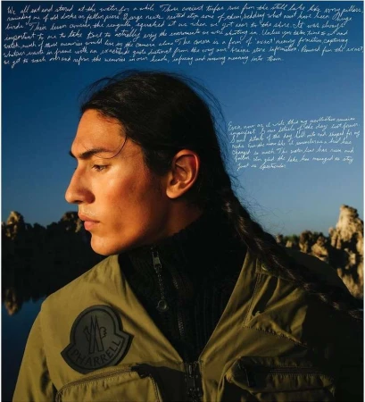 Profile portrait of an adult male with long braided hair, wearing olive green outerwear in an outdoor lakeside setting with rock formations.