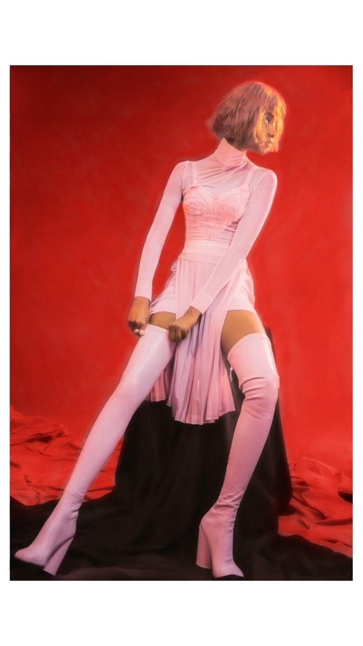 Fashion photo of a woman wearing a pink dress with thigh-high boots sitting on a black draped surface in front of a red background.