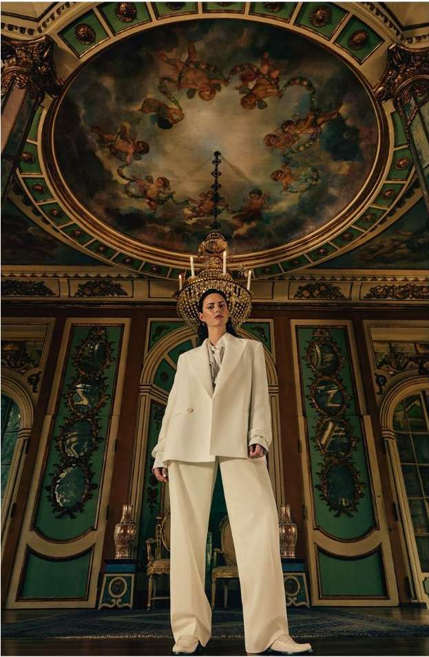 A woman in a white pantsuit stands in an ornate room with detailed wall panels and a ceiling fresco with a chandelier overhead.
