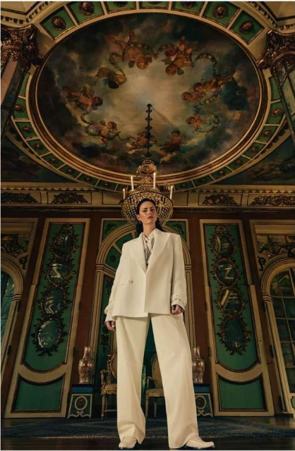 A woman in a white pantsuit stands in an ornate room with detailed wall panels and a ceiling fresco with a chandelier overhead.
