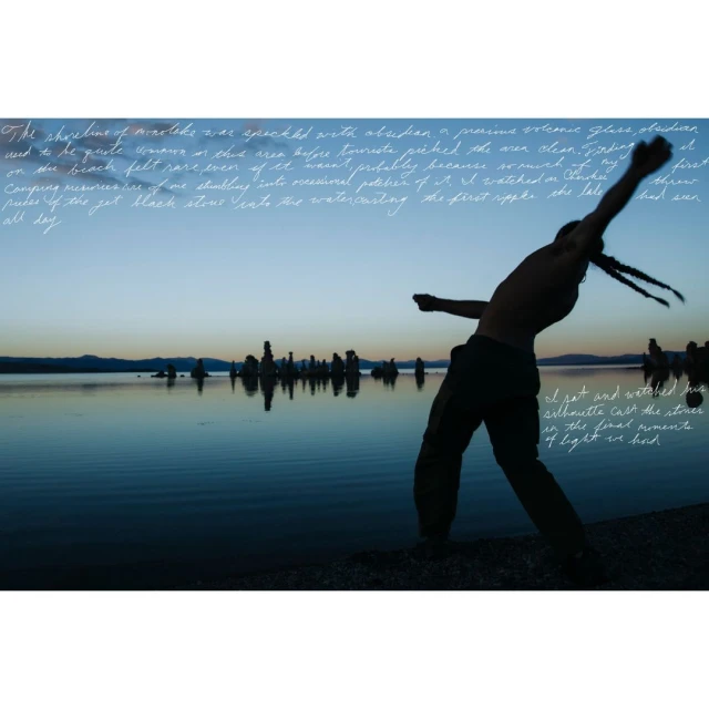 Silhouette of shirtless adult male throwing a stone into calm lake at sunset near rock formations; blue and orange hues dominate the scene.