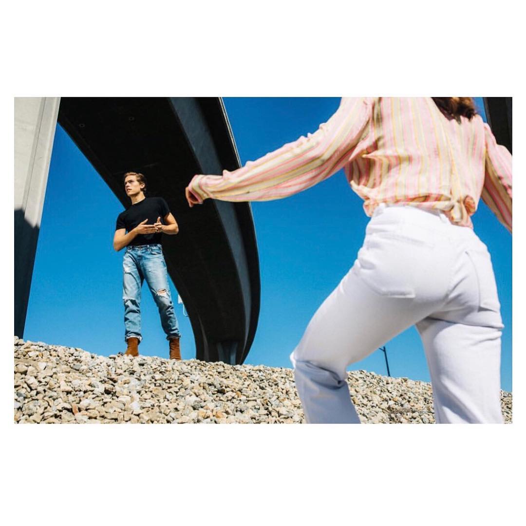 Two people posing outdoors on a rocky surface beneath a concrete structure, with a clear blue sky in the background and wearing casual clothing.
