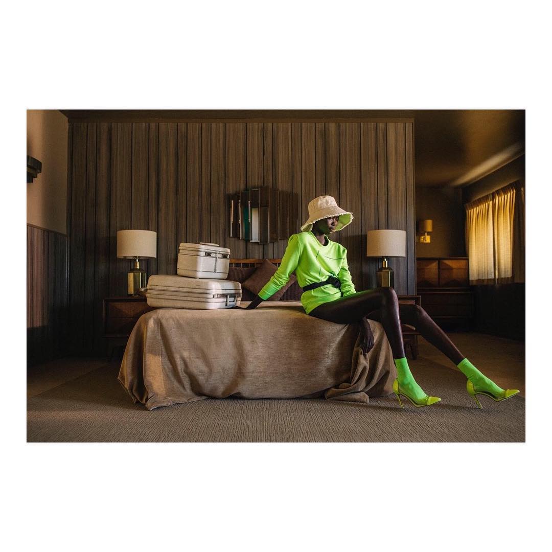 A fashion shot of a woman in a neon green outfit and hat sitting on a bed with vintage suitcases in a neutral-toned hotel room.