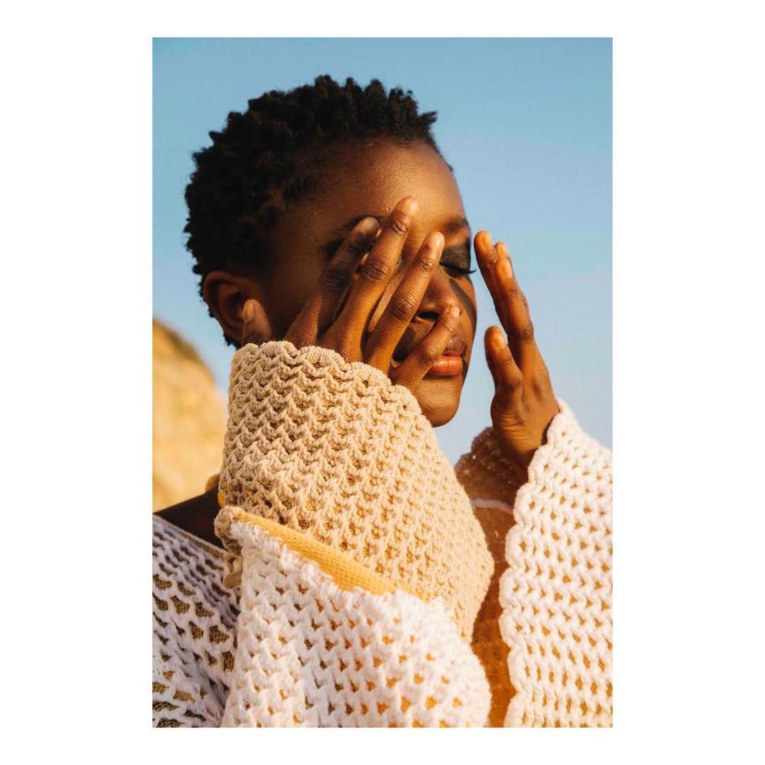 Close-up of a stylish African woman with short hair covering her face with her hands, wearing a textured beige sweater against a blue sky.