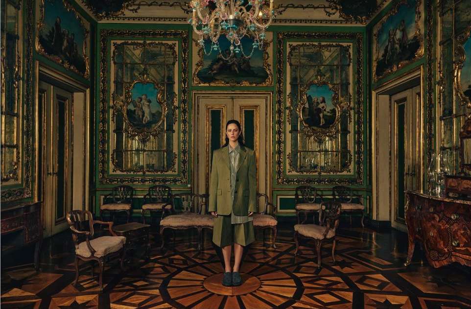 Adult female stands centered in a vintage hall wearing a green blazer and shorts, with ornate details, chandelier, and parquet floor.
