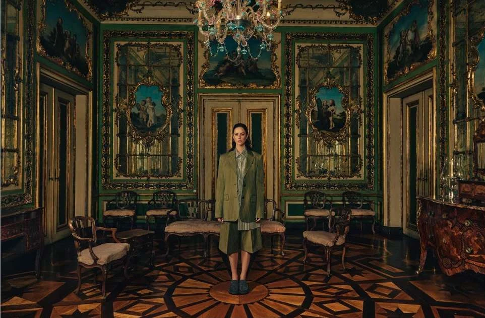 Adult female stands centered in a vintage hall wearing a green blazer and shorts, with ornate details, chandelier, and parquet floor.