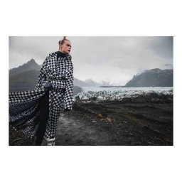 Model wearing a black and white houndstooth coat standing in a rugged mountain landscape with glaciers and snowy peaks under an overcast sky.