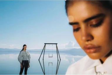 Close-up of a woman in the foreground with another figure standing near a swing on a reflective shallow body of water under a clear blue sky