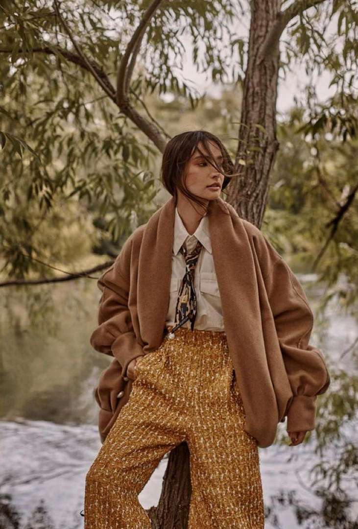A woman in fashionable outfit poses near a tree in an outdoor setting, wearing a tan jacket, patterned top, and gold-colored pants.