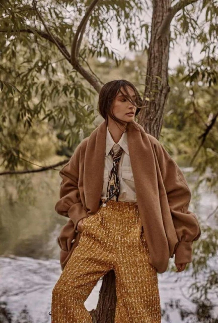 A woman in fashionable outfit poses near a tree in an outdoor setting, wearing a tan jacket, patterned top, and gold-colored pants.