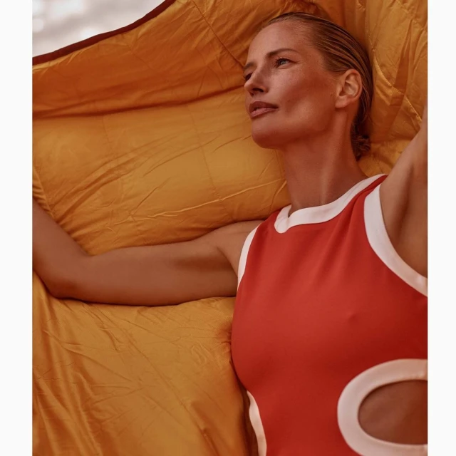 Adult woman poses wearing a one-piece swimsuit, draped with a mustard-yellow quilted blanket; the shot emphasizes her natural appearance.
