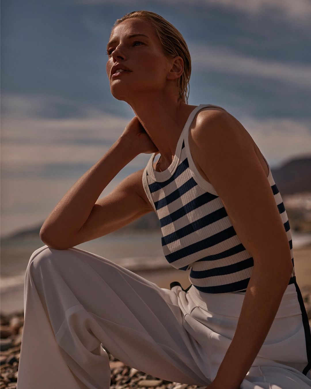 Woman in striped tank top and white trousers squatting on a beach with mountains in the background under an overcast sky.