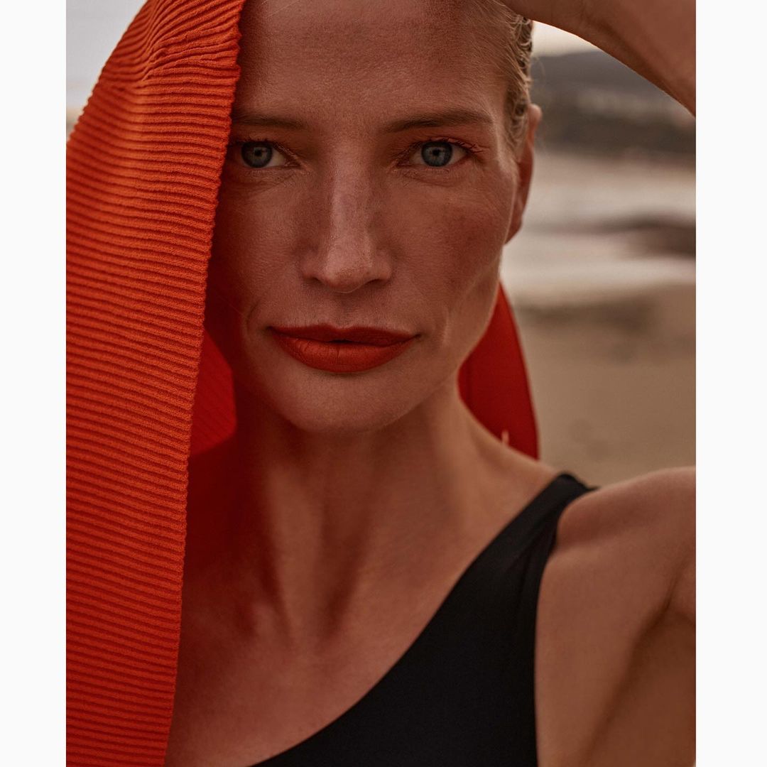 Close-up portrait of a woman with an orange pleated fabric draped over her head and shoulders, wearing a black swimsuit on a beach.