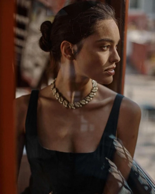 A portrait of a young woman with dark hair and a black dress looking out of a window, wearing a leaf-patterned necklace. Natural lighting.