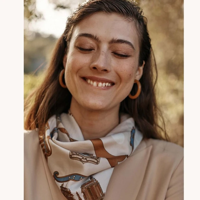 Portrait of a smiling woman with closed eyes wearing a printed scarf and blazer in an outdoor setting with blurred background.