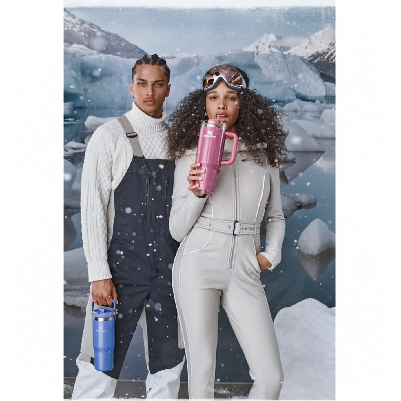 Two people, a man and a woman, pose in a snowy landscape wearing winter clothing and holding Stanley mugs, creating a cold and stylish scene.