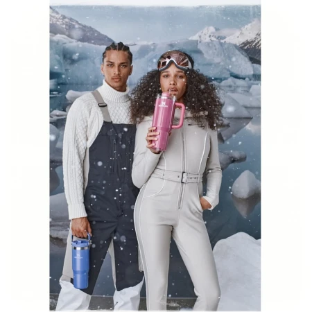 Two people, a man and a woman, pose in a snowy landscape wearing winter clothing and holding Stanley mugs, creating a cold and stylish scene.