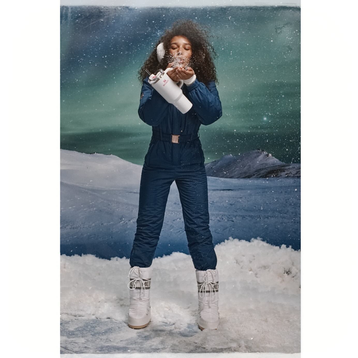 Young woman in snowsuit and boots playfully blowing snow in a wintry landscape, wearing earmuffs with a whimsical, snowy background.
