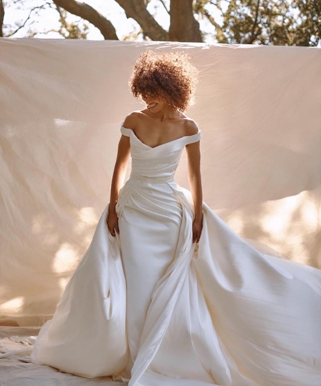 Elegant woman in a white wedding dress posing outside with an off-shoulder neckline, a sweeping train, and a neutral backdrop.