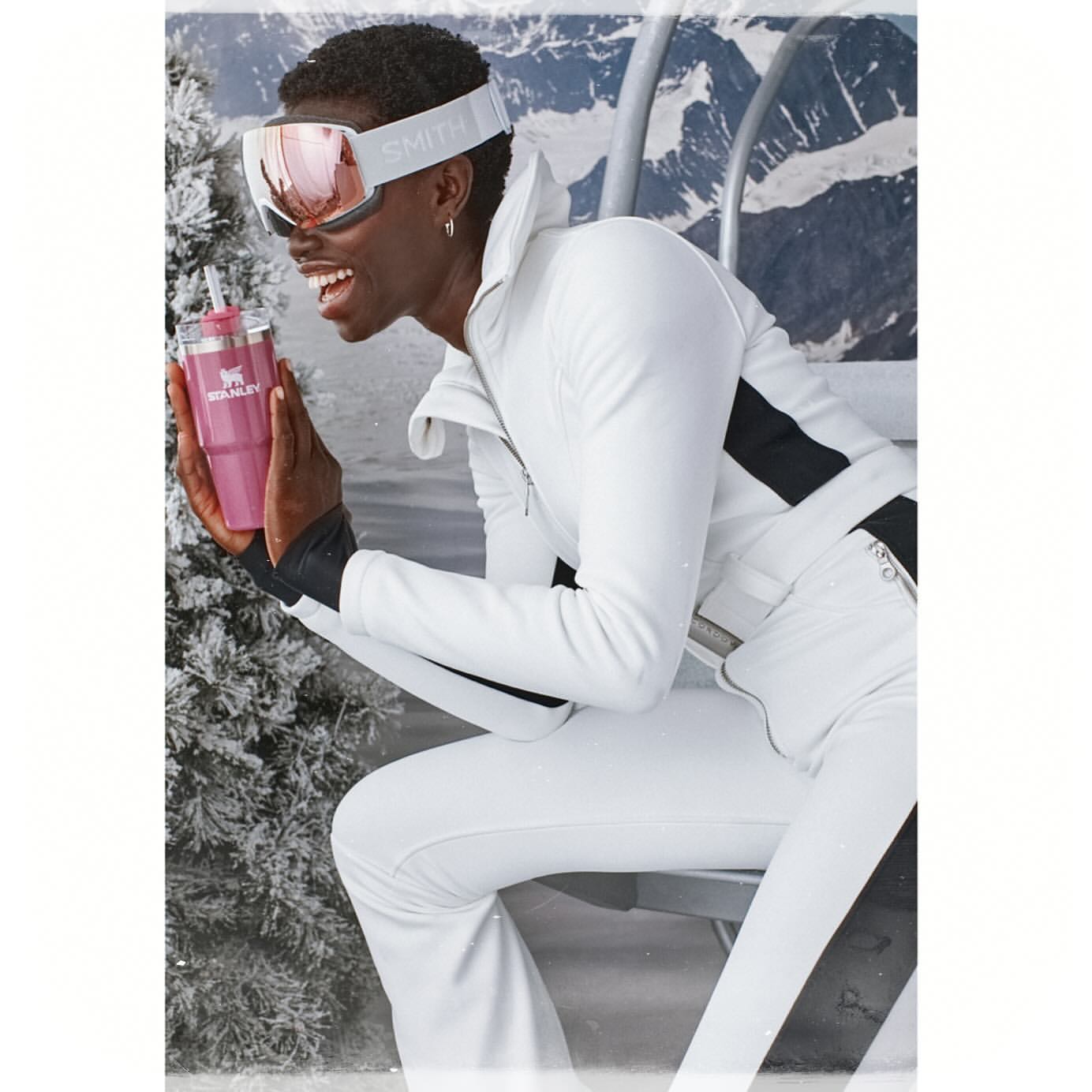 A smiling woman in ski apparel sits on a lift, holding a pink Stanley tumbler, with a snow-covered mountain backdrop.