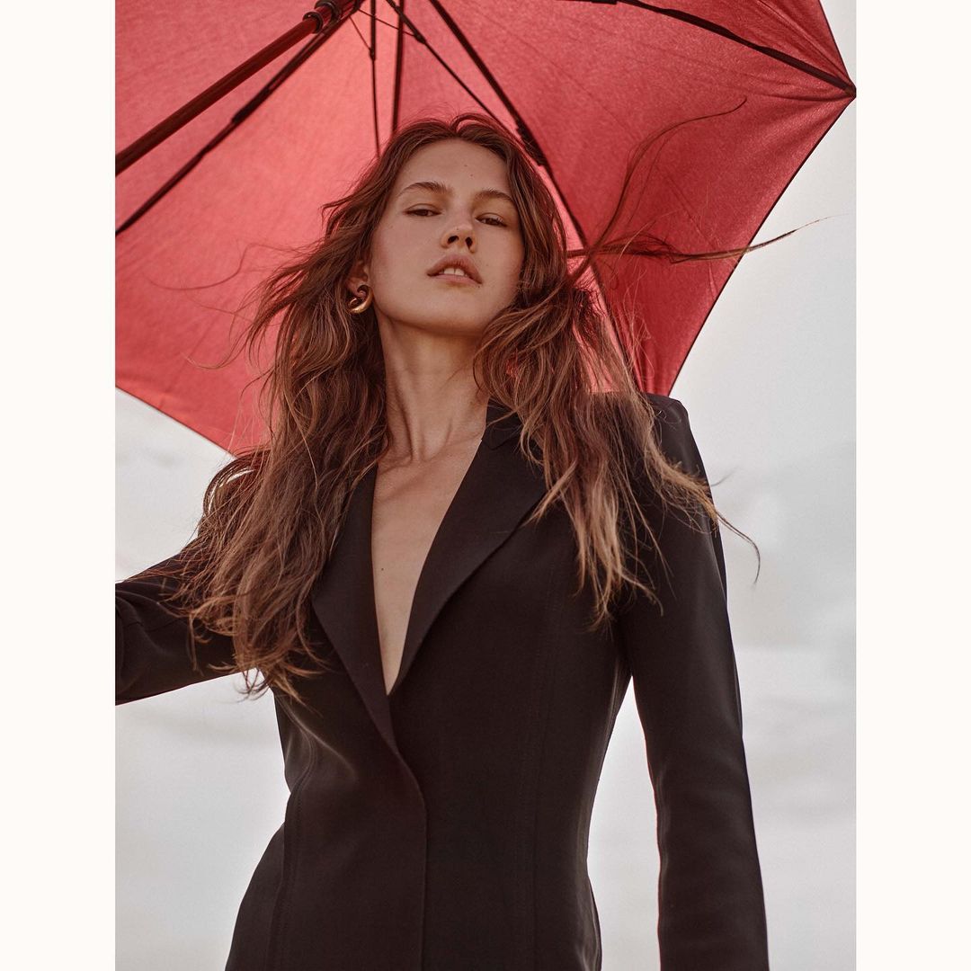 Woman in a black jacket with long hair stands with a red umbrella against a white and tan sky in a medium portrait shot.