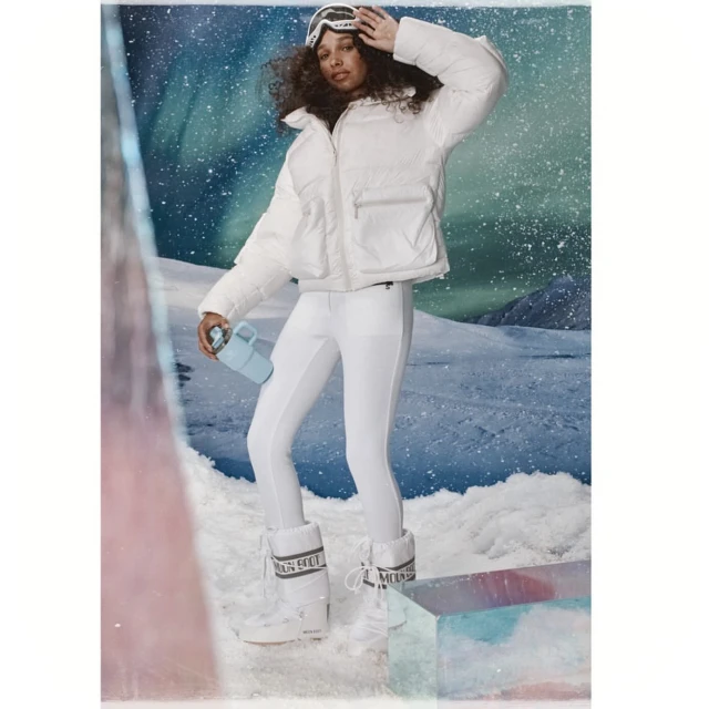 A woman poses outdoors in snow wearing a white puffer jacket, leggings, and moon boots, holding a blue bottle in a winter fashion shoot.
