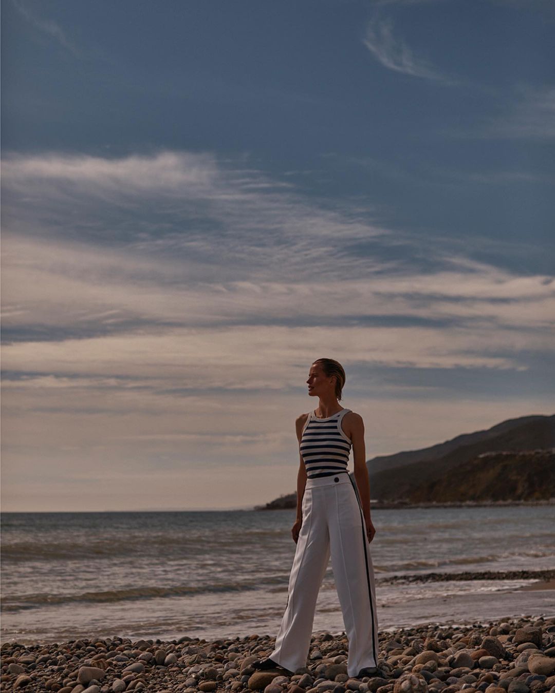 A woman stands on a pebble beach wearing a striped top and white pants with the ocean and distant shoreline in the background.