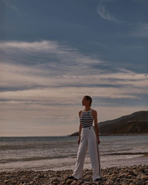 A woman stands on a pebble beach wearing a striped top and white pants with the ocean and distant shoreline in the background.