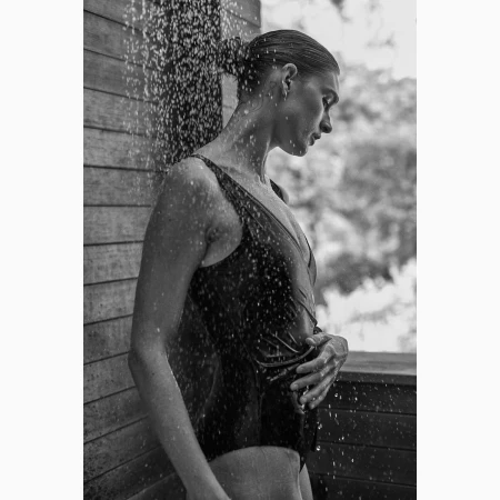 Black and white portrait of a woman standing outdoors under falling water in a swimsuit, photographed by Colette De Barros.