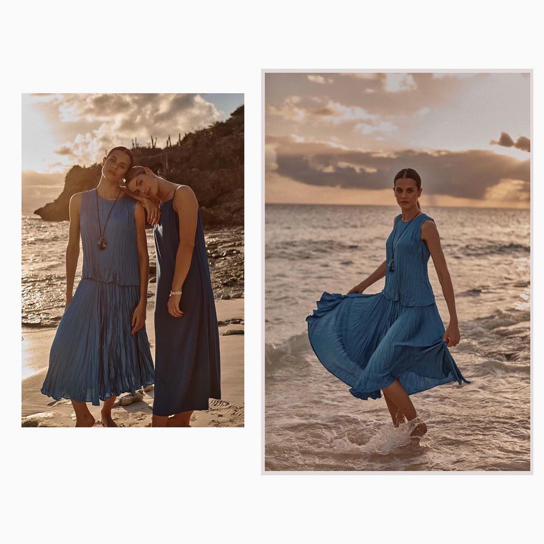 Two women in pleated dresses pose on a beach next to a woman wading into the ocean in a turquoise dress at sunset.