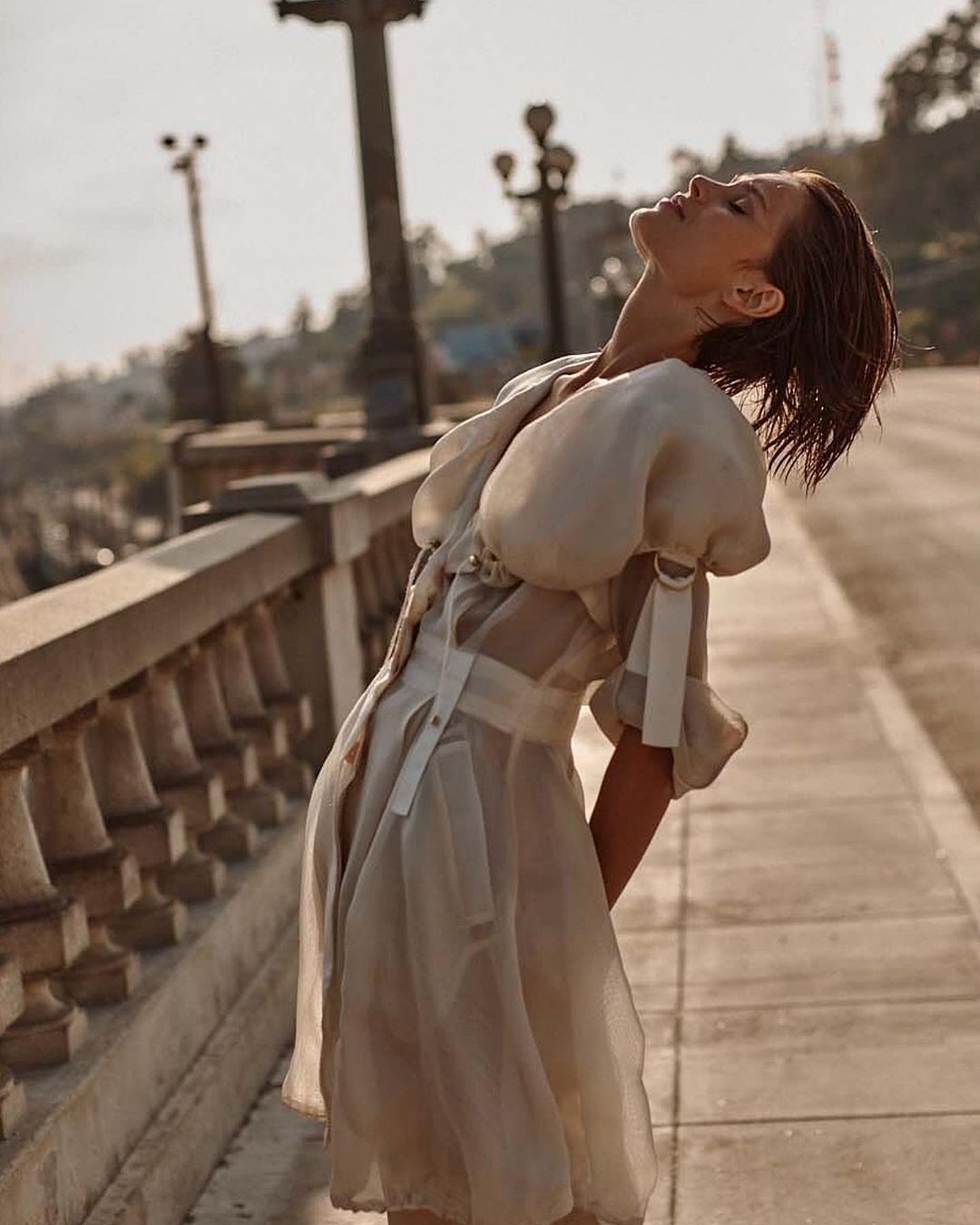 Woman in flowing beige dress with puffed sleeves standing on a stone bridge enjoying the warm sun of a clear day.